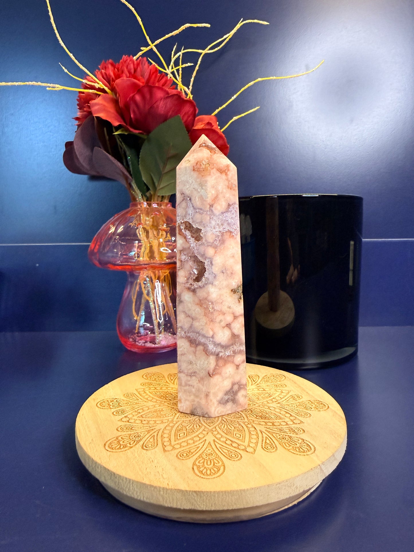A pink and peach crystal tower with druzy pockets on a decorative coaster in front of a red flower and black jar