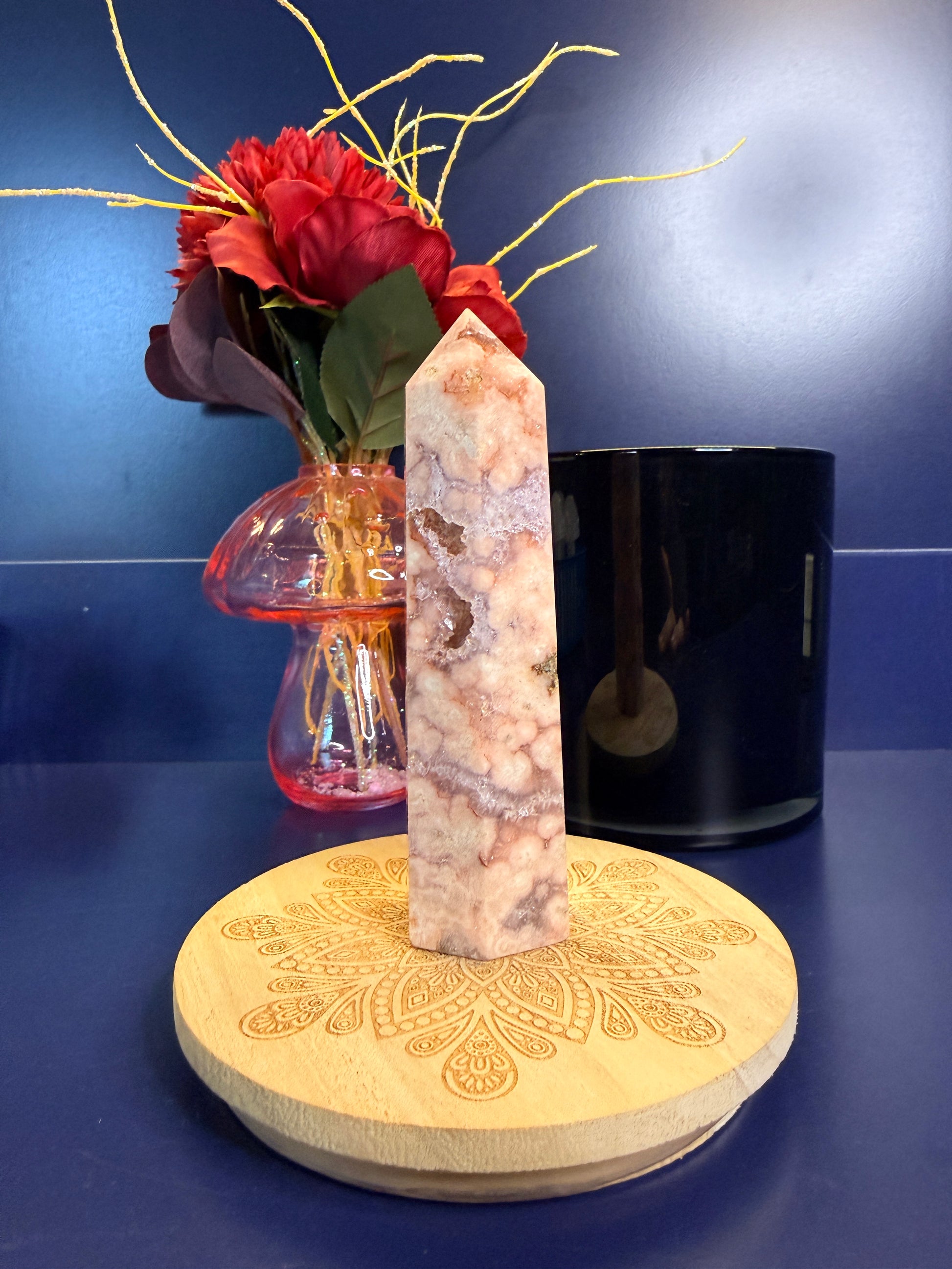 A pink and peach crystal tower with druzy pockets on a decorative coaster in front of a red flower and black jar