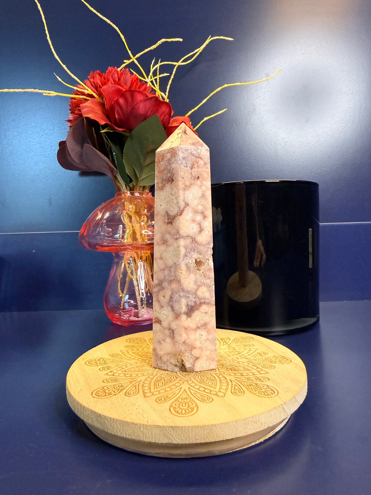 A pink and peach crystal tower with druzy pockets on a decorative coaster in front of a red flower and black jar