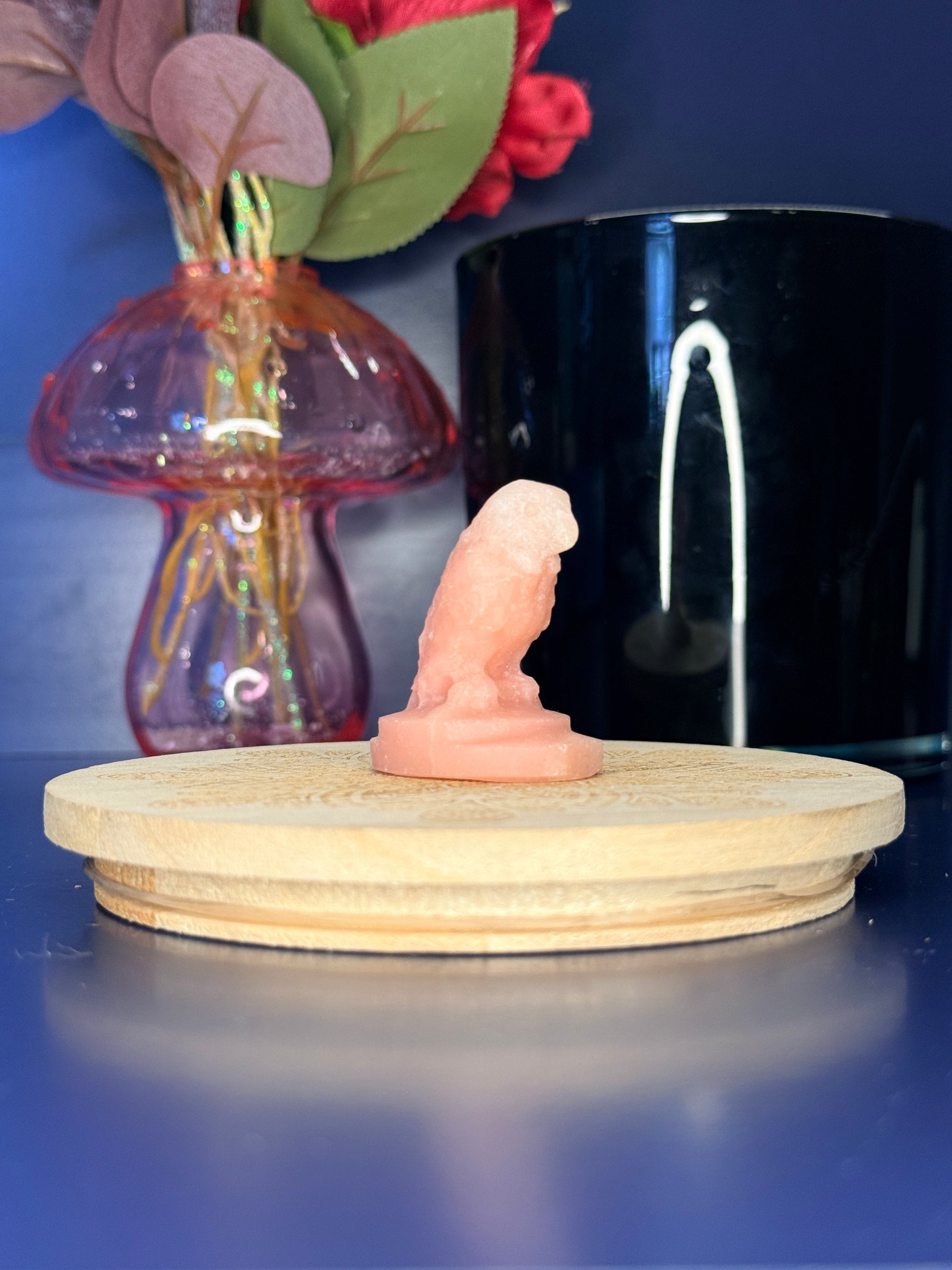 Pink opal parrot carving in front of a pink vase and black jar on a wooden coaster against a blue background