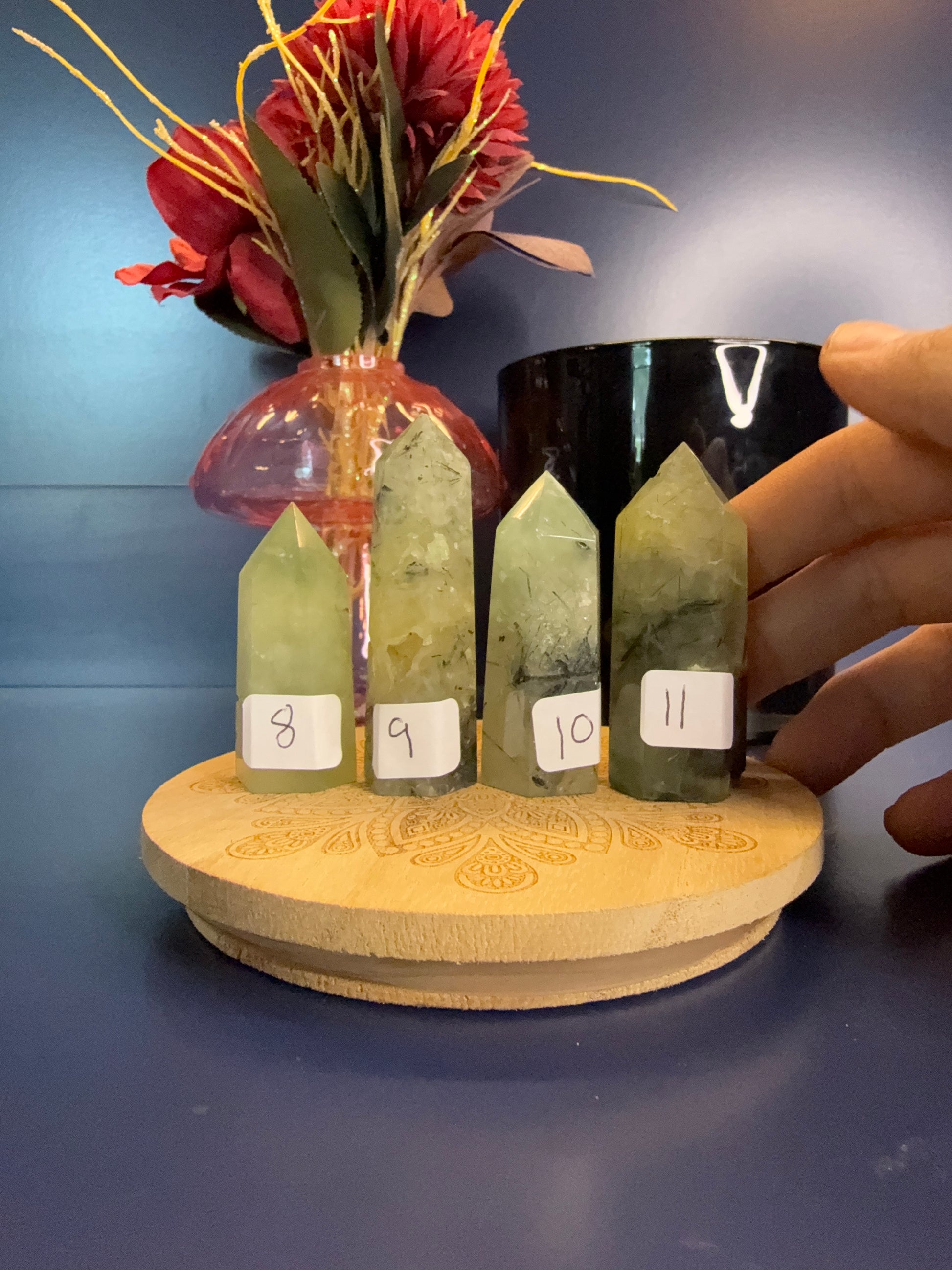 Four light green and black prehnite towers in front of a red flower and black jar, with a hand in the right side of the frame