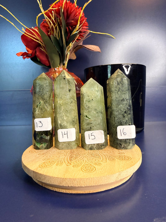 Four light green and black prehnite towers in front of a red flower and black jar on a wooden coaster