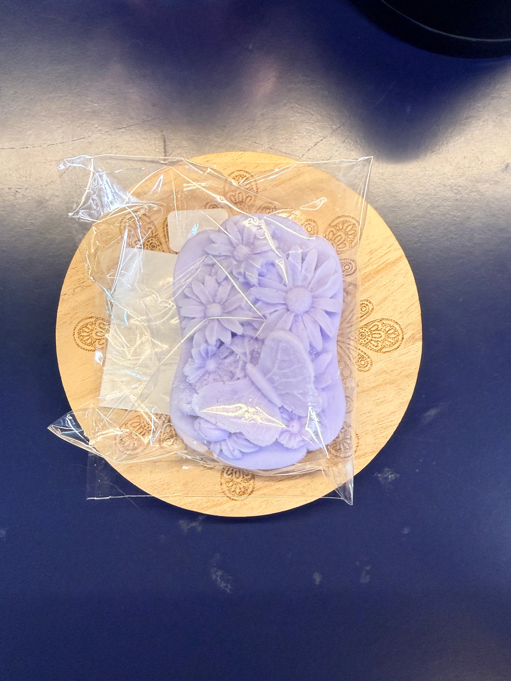 Purple soap bar with a decorative design on a wooden coaster, wrapped in clear plastic on a dark surface.