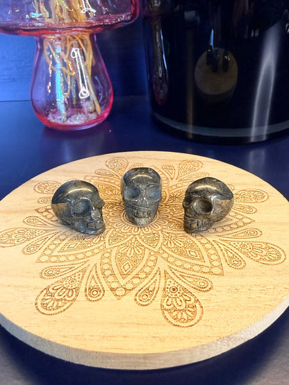 Three dark gold pyrite skulls on a decorative wooden coaster in front of a vase and black jar