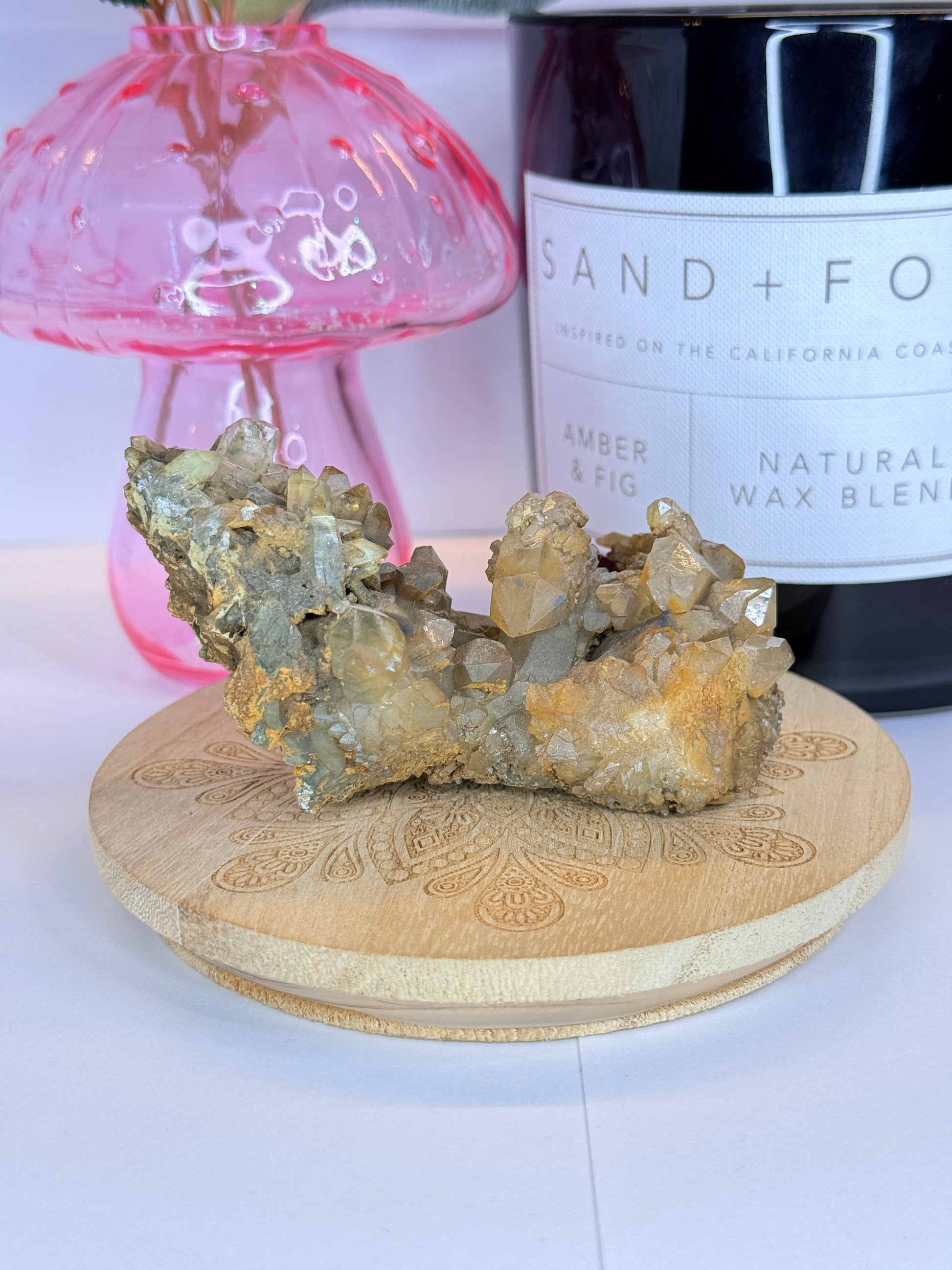 Unique quartz and hematite specimen resting on a wooden coaster in front of a black candle jar and pink vase