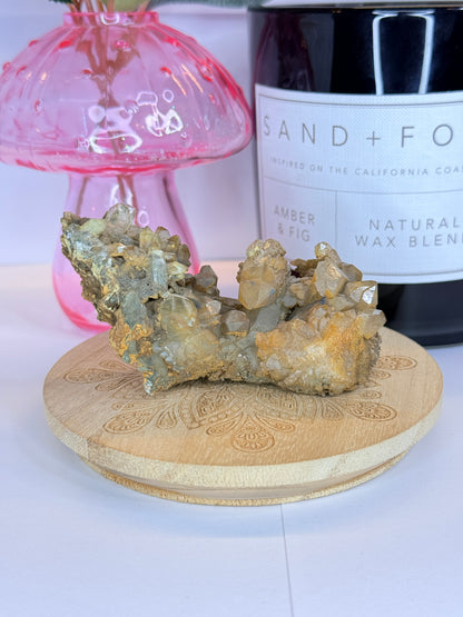 Unique quartz and hematite specimen resting on a wooden coaster in front of a black candle jar and pink vase