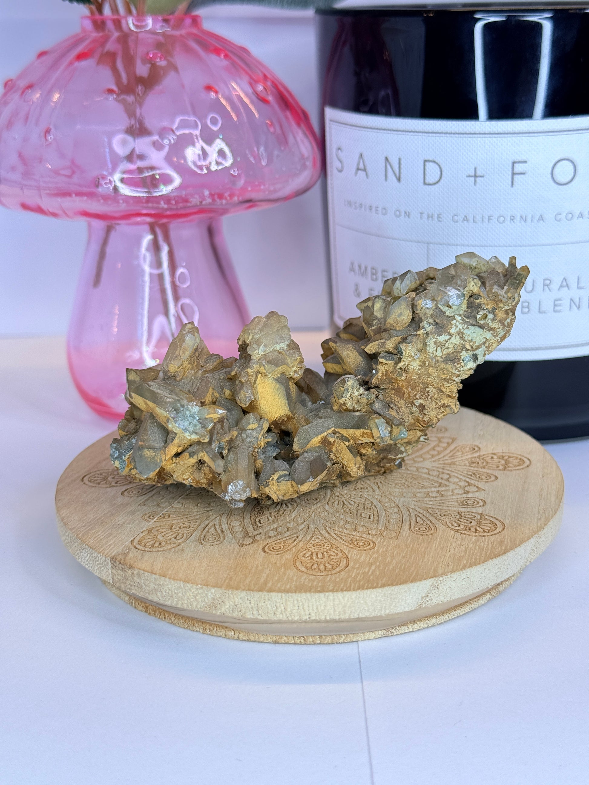 Unique quartz and hematite specimen resting on a wooden coaster in front of a black candle jar and pink vase