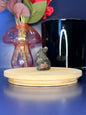 Blue and black que sera bunny carving in front of a pink vase and black jar on a wooden coaster against a blue background