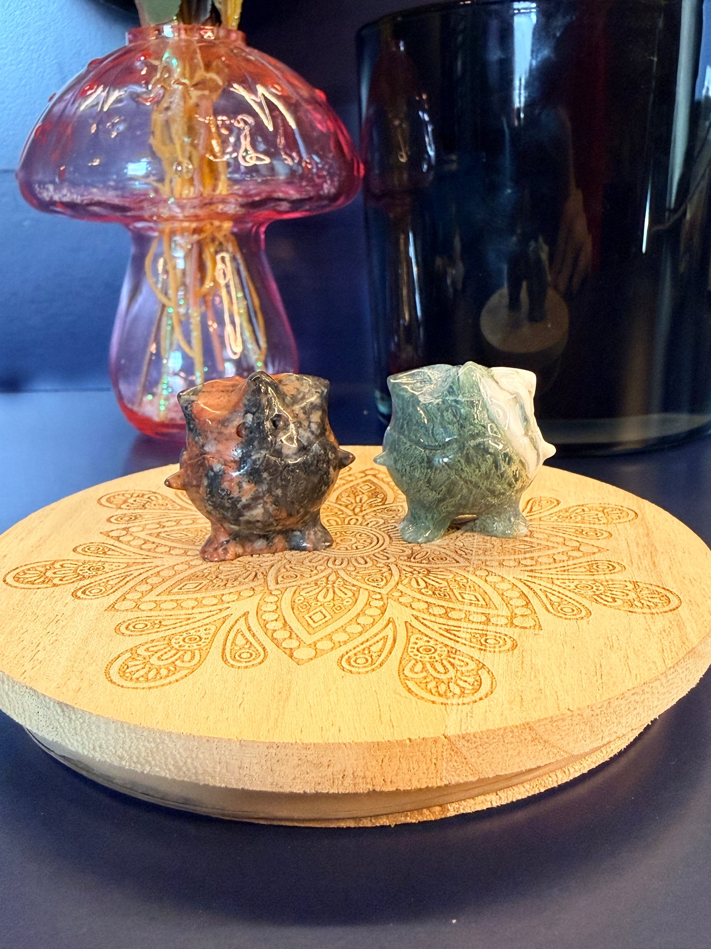 Two Togepi-inspired shapes, que sera and moss agate, on a wooden coaster in front of a glass mushroom and black jar