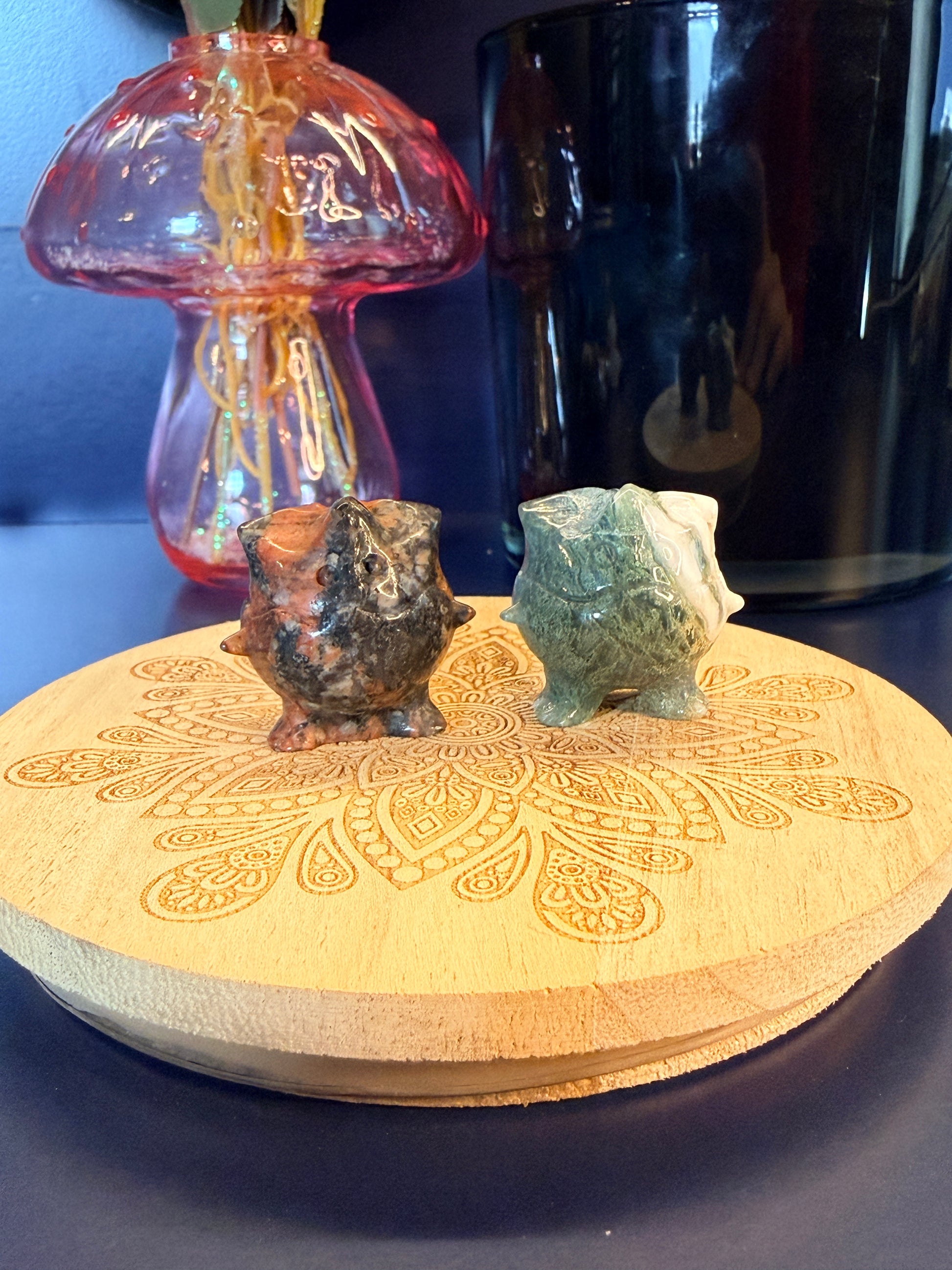 Two Togepi-inspired shapes, que sera and moss agate, on a wooden coaster in front of a glass mushroom and black jar