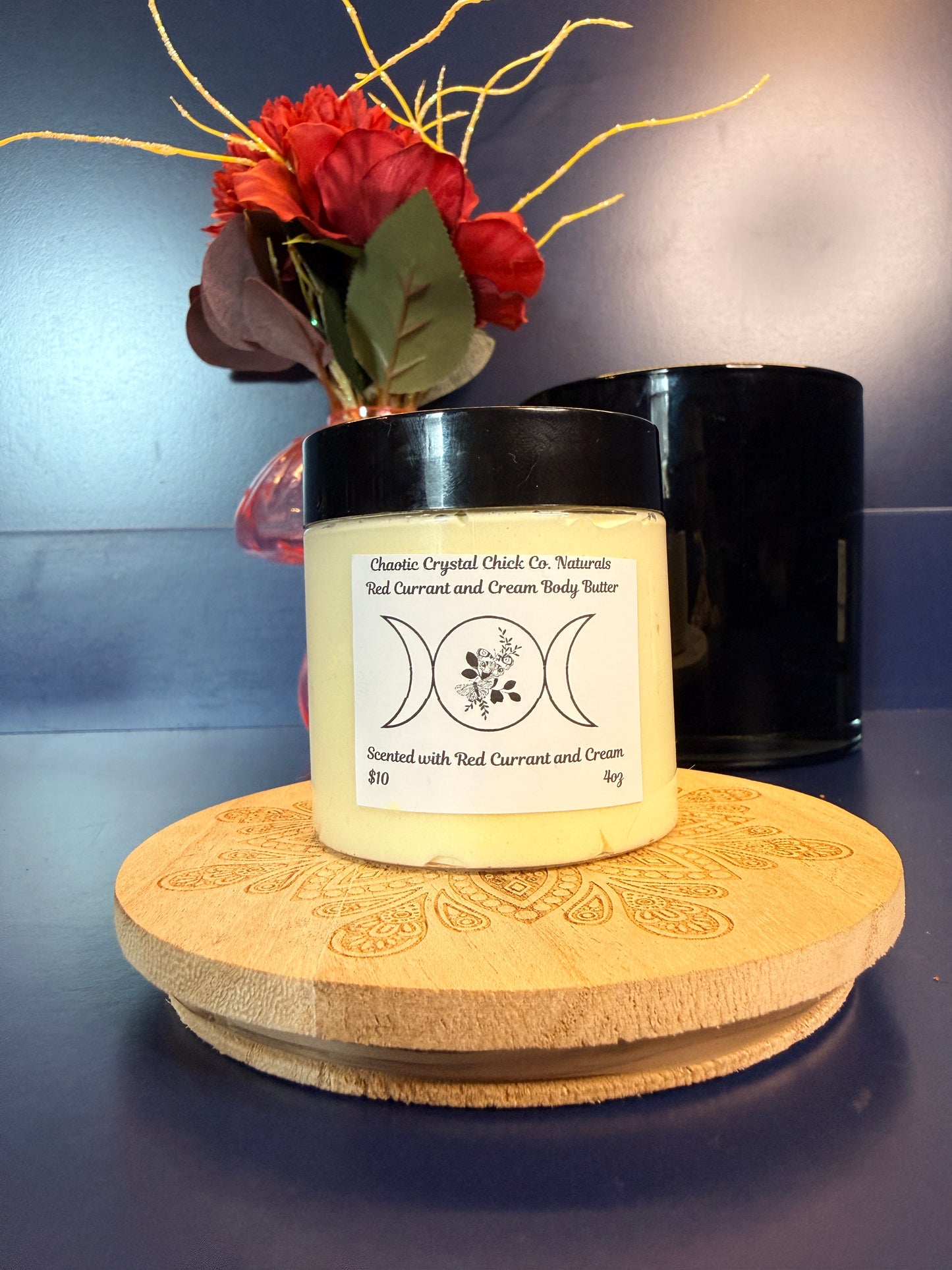 Jar of yellow body butter with a label on a decorative wooden coaster in front of a red flower and black jar