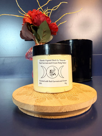 Jar of yellow body butter with a label on a decorative wooden coaster in front of a red flower and black jar