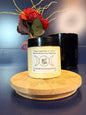 Jar of yellow body butter with a label on a decorative wooden coaster in front of a red flower and black jar