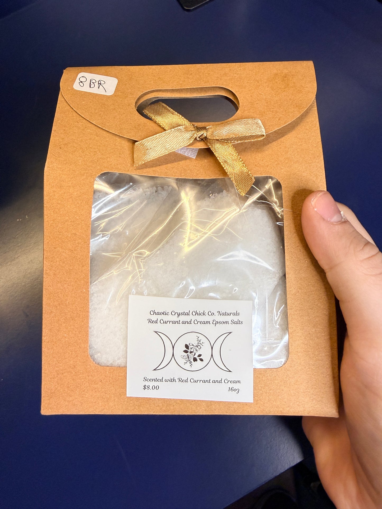 Hand holding white epsom salt in a brown paper container with a bow on a blue background