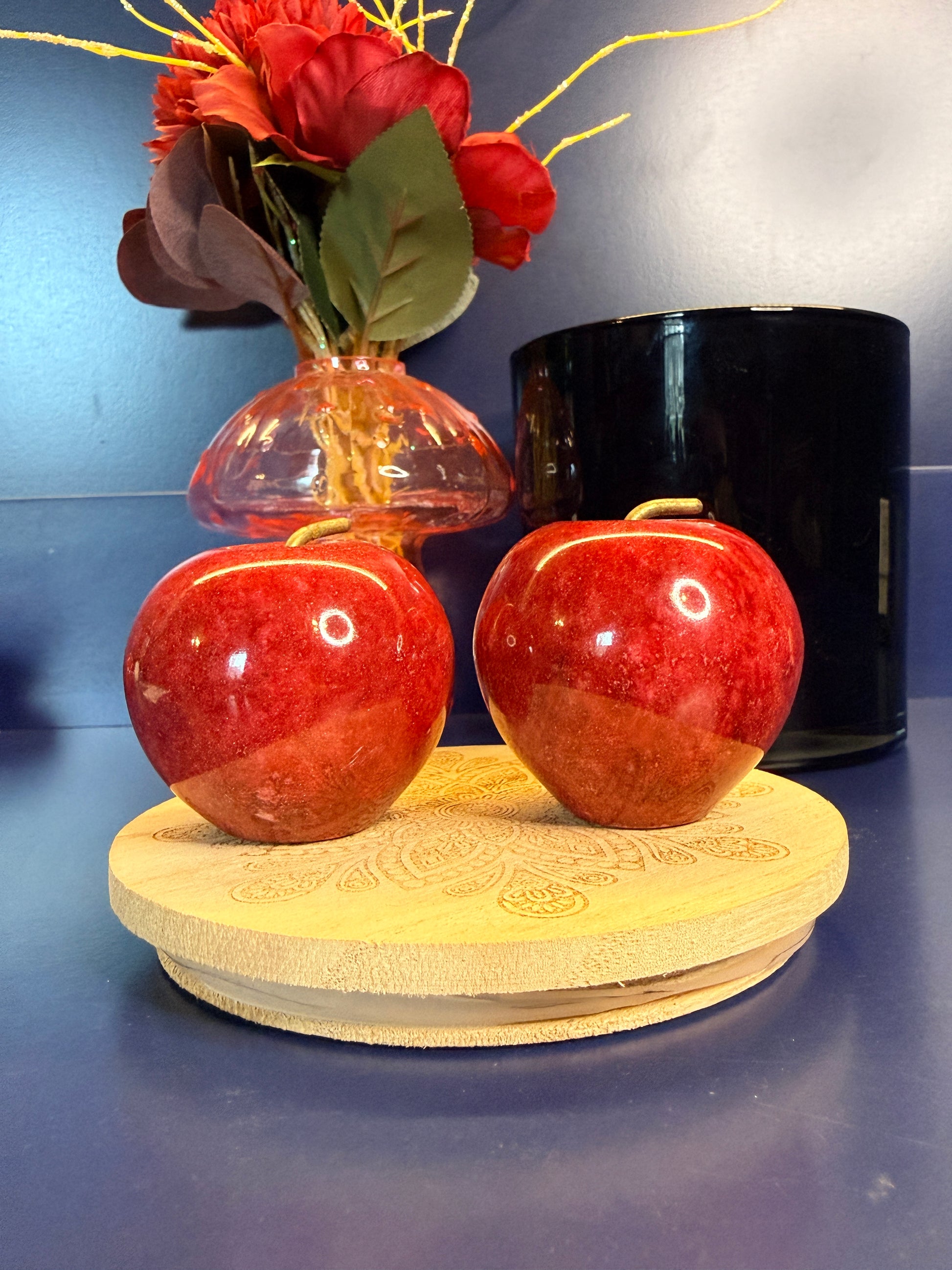 Two red jasper apples on a decorative wooden coaster in front of red flowers in a mushroom vase and black jar