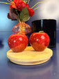 Two red jasper apples on a decorative wooden coaster in front of red flowers in a mushroom vase and black jar