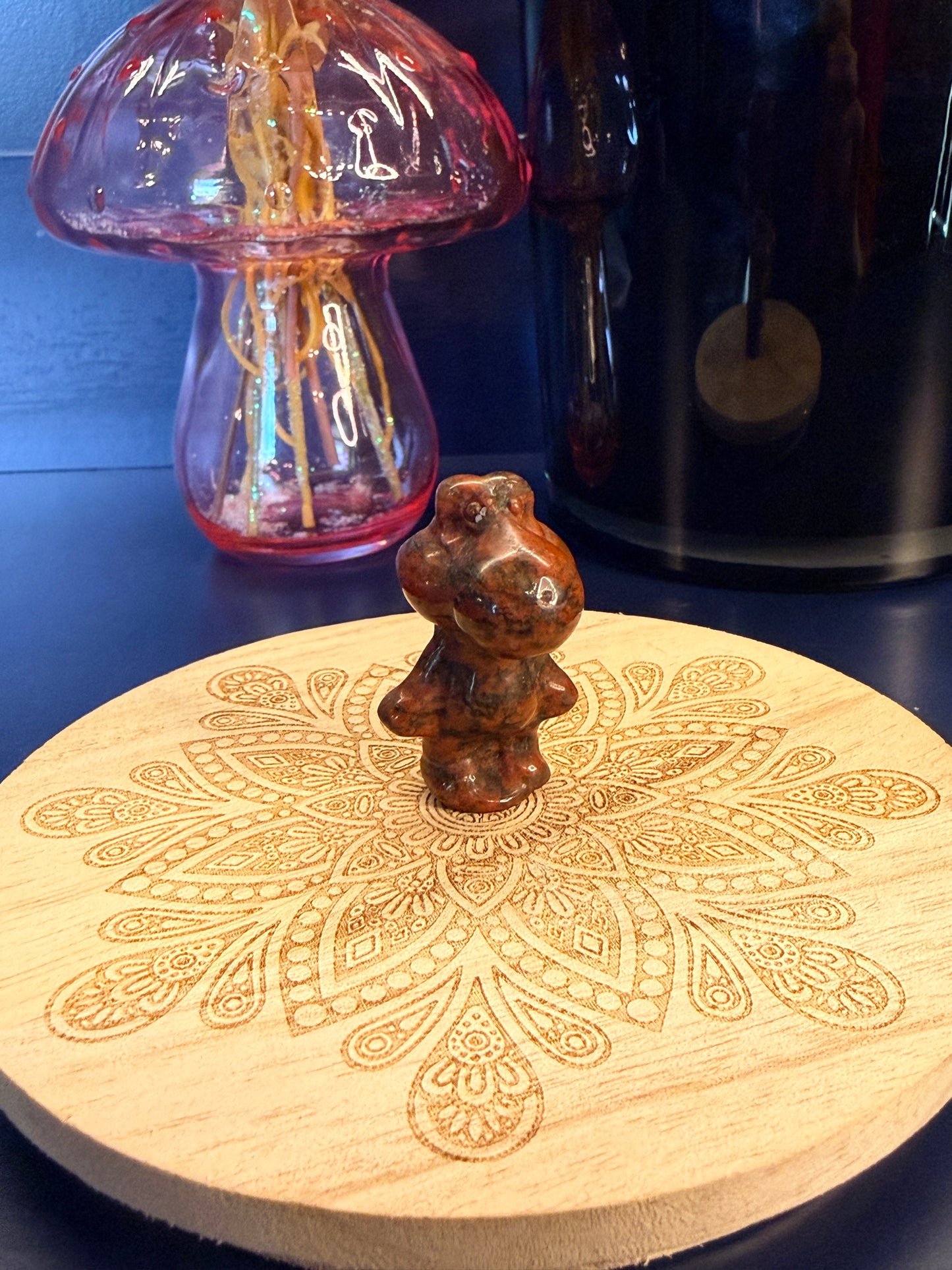 A red sesame jasper yoshi crystal sitting on a decorative wooden coaster in front of a glass mushroom and black jar
