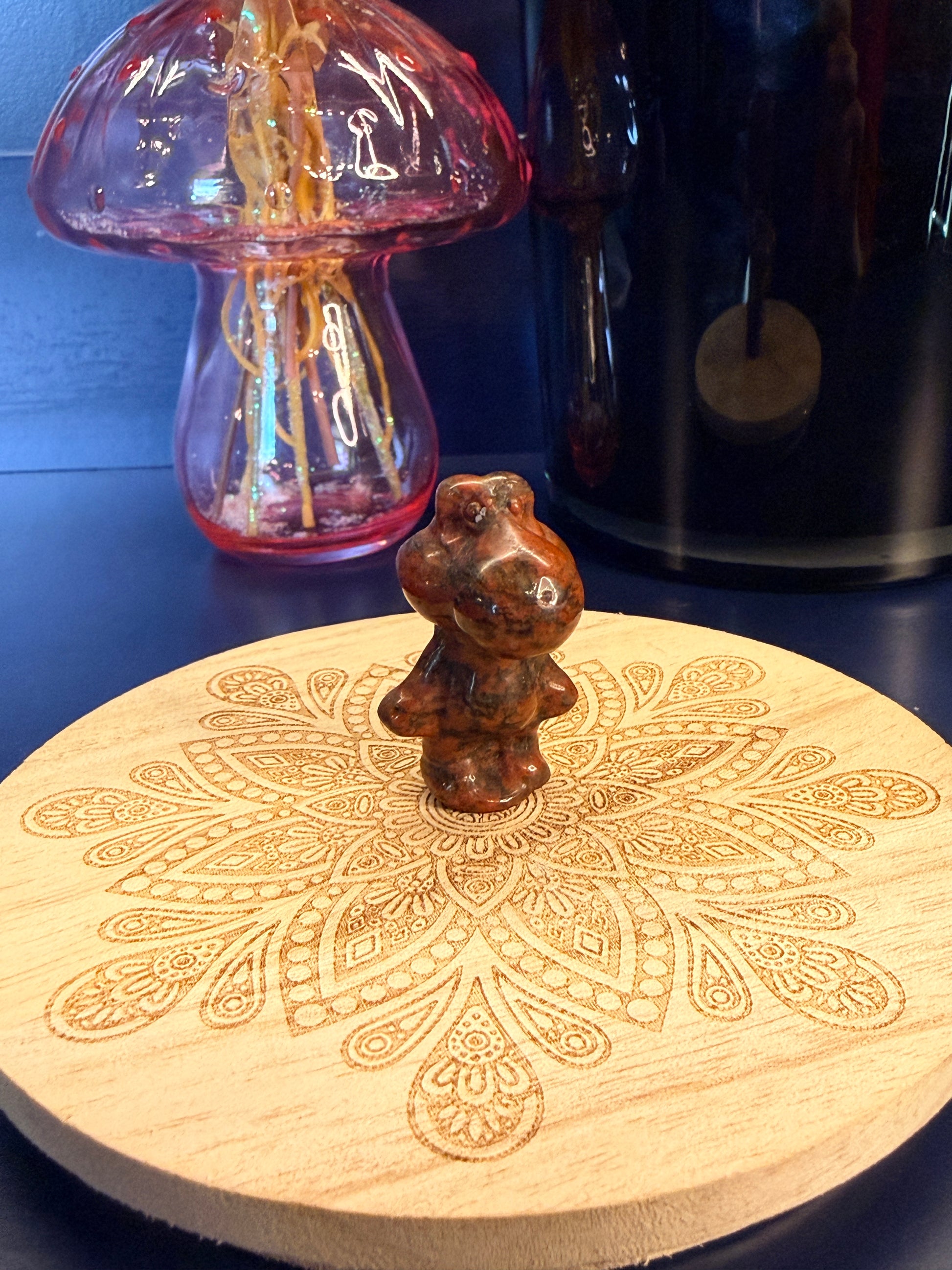 A red sesame jasper yoshi crystal sitting on a decorative wooden coaster in front of a glass mushroom and black jar