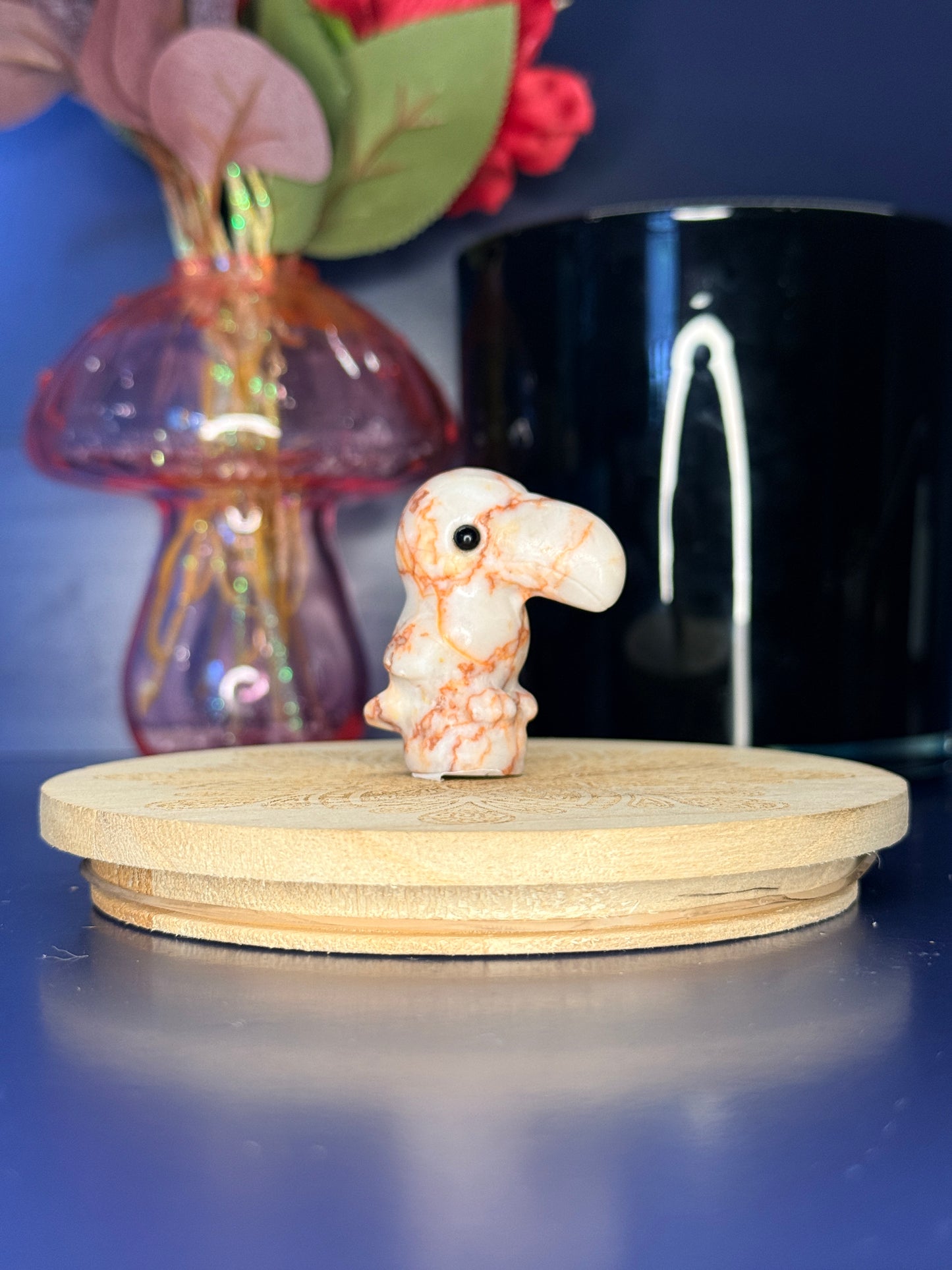 Red and white jasper toucan carving in front of a pink vase and black jar on a wooden coaster against a blue background