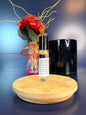 Body Oil Vial sitting on a decorative wooden coaster in front of a red flower and black jar