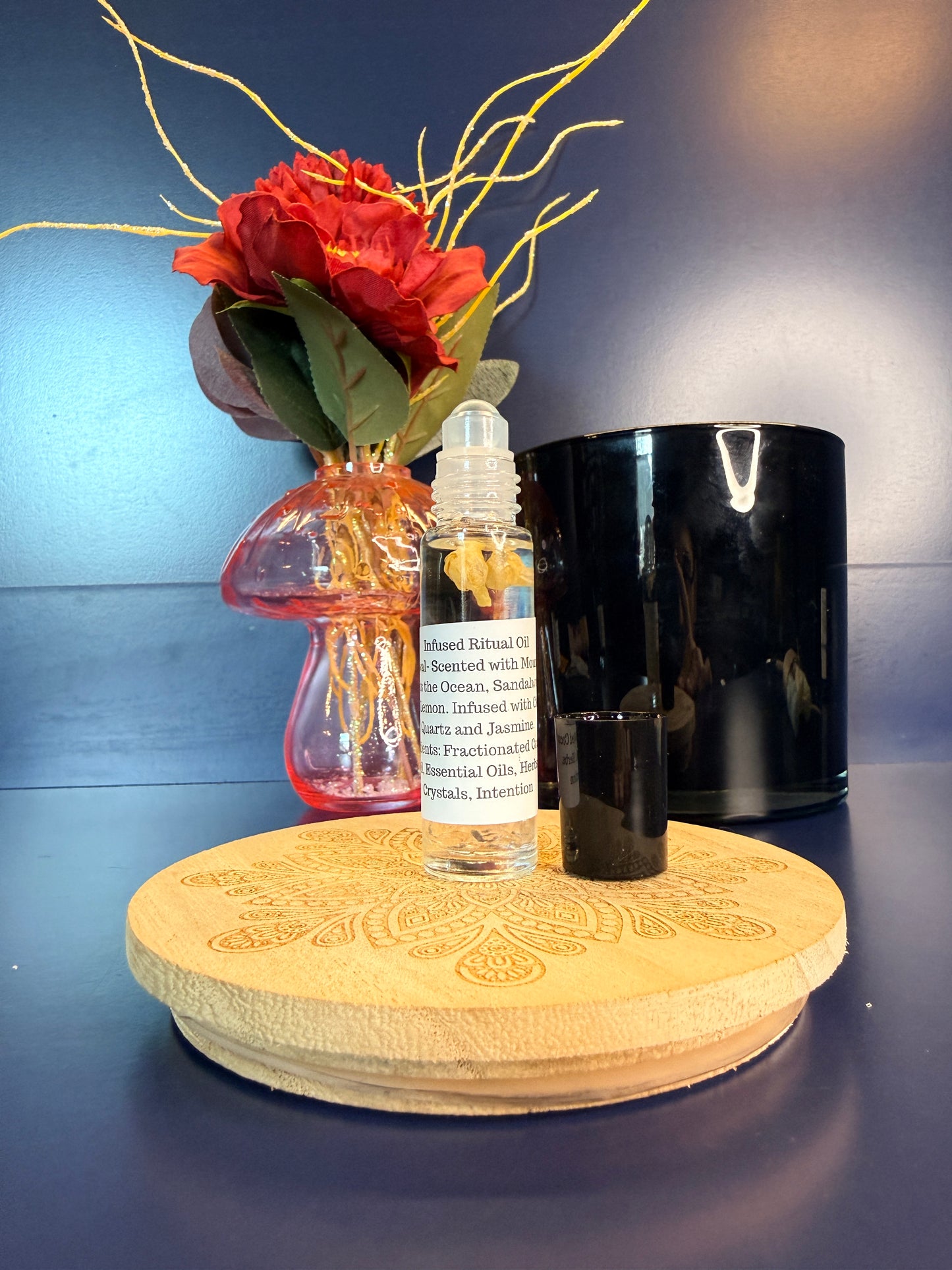 Body Oil Vial sitting on a decorative wooden coaster in front of a red flower and black jar