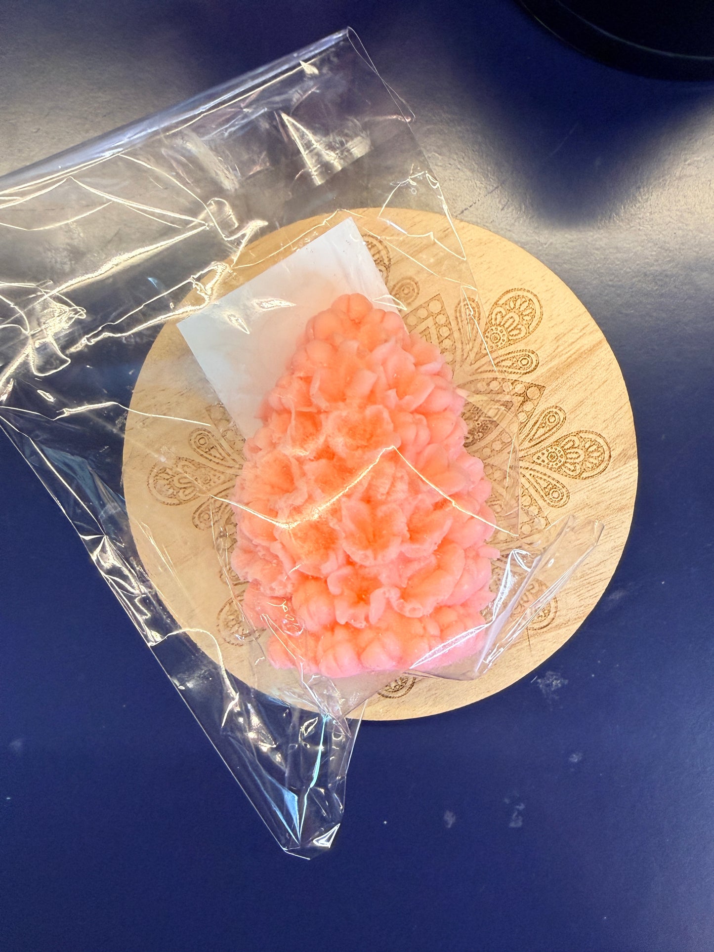 Orange flower-shaped soap on a decorative wooden coaster with clear plastic wrap.