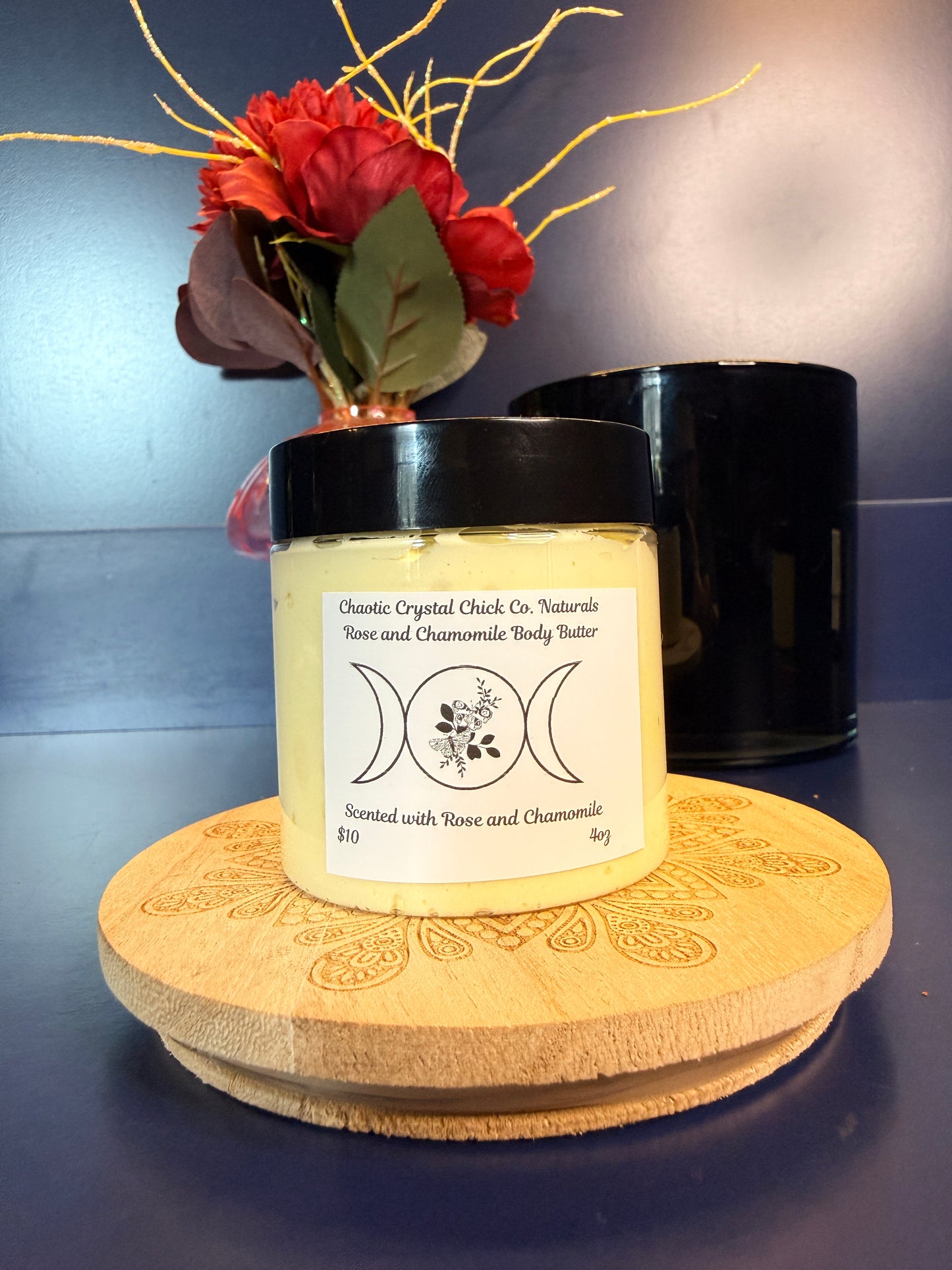 Jar of yellow body butter with a label on a decorative wooden coaster in front of a red flower and black jar