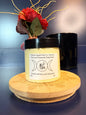Jar of yellow body butter with a label on a decorative wooden coaster in front of a red flower and black jar