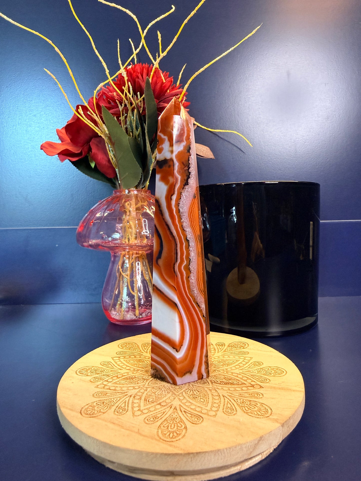 A black, white, and red sardonyx tower sitting on a decorative wooden coaster in front of a mushroom vase and black jar