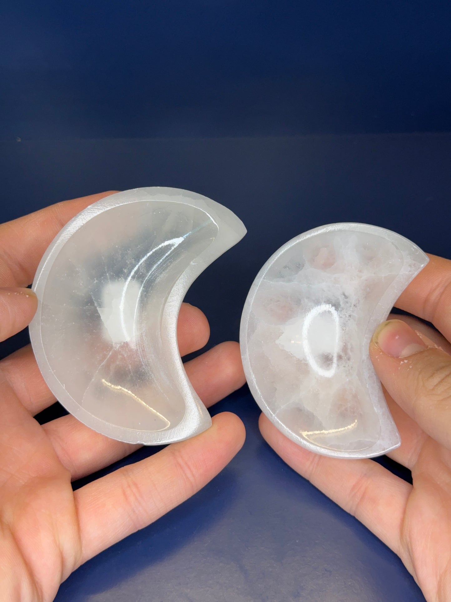 Two hands holding white selenite moon bowls on a blue background