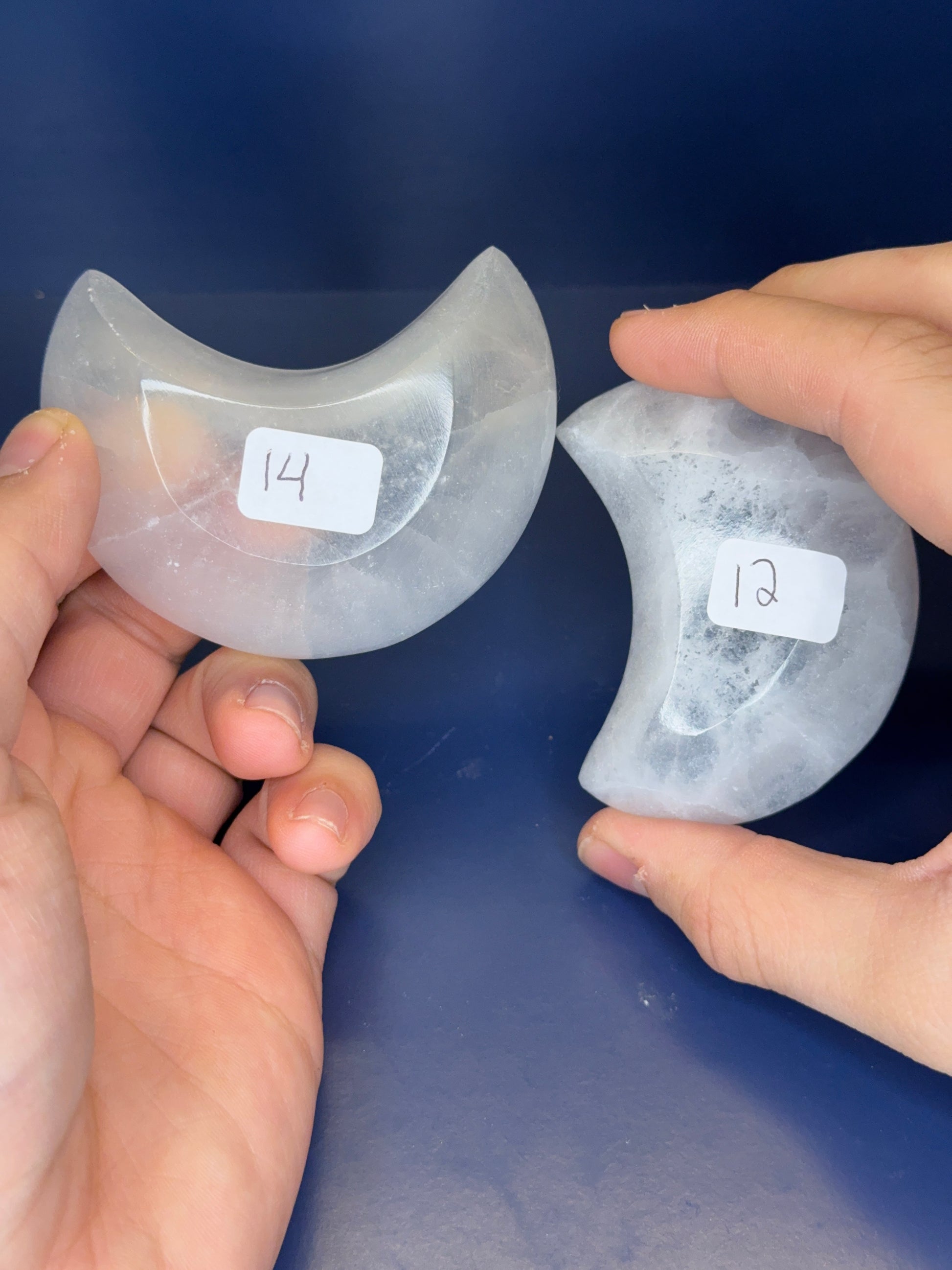 Two hands holding white selenite moon bowls with labels on a blue background