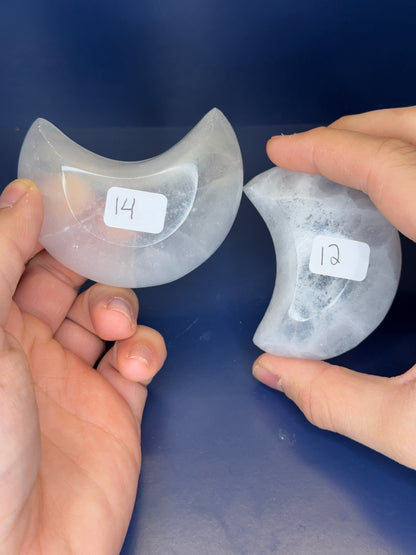 Two hands holding white selenite moon bowls with labels on a blue background