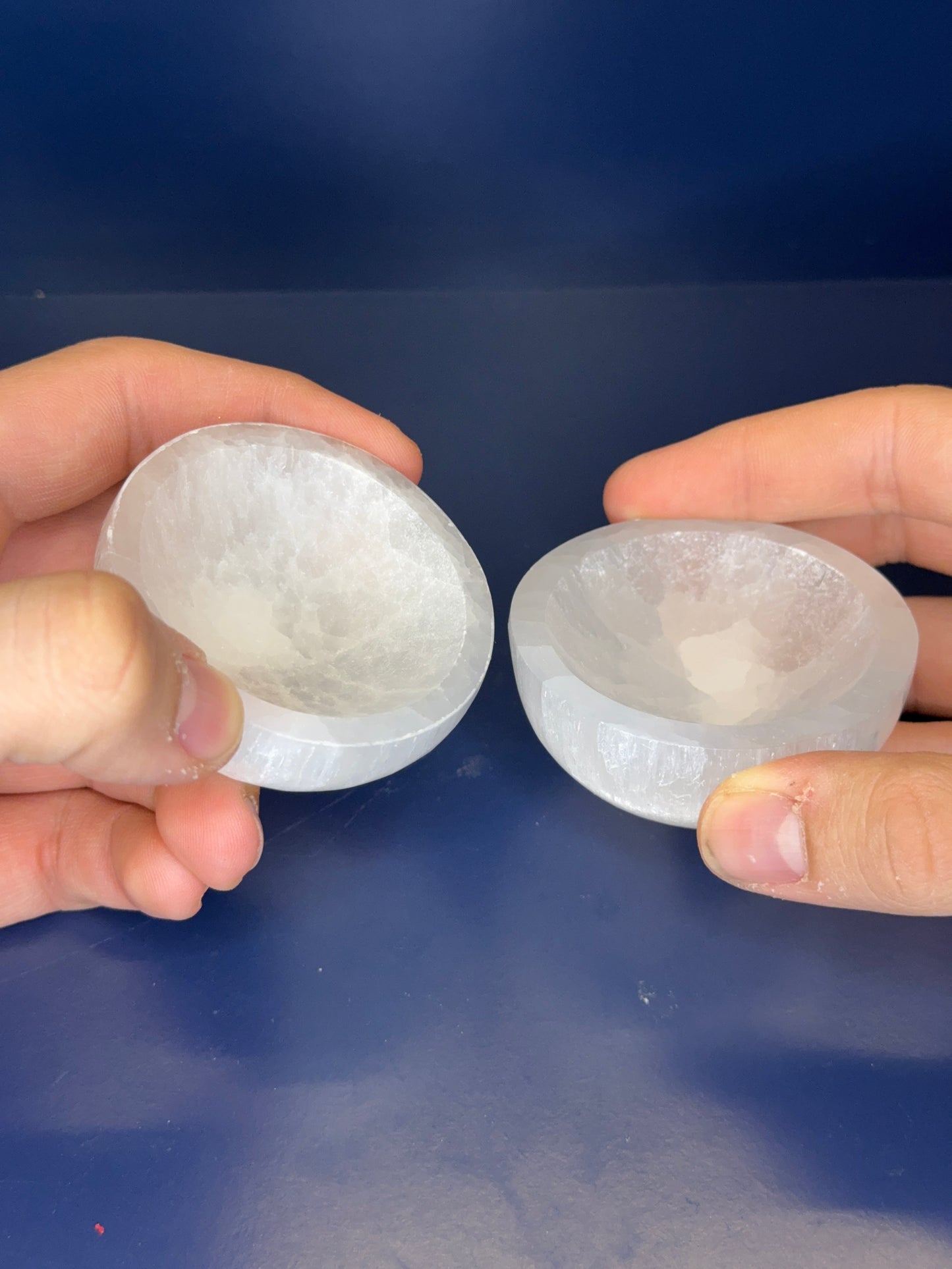 Two hands holding white selenite bowls on a blue background