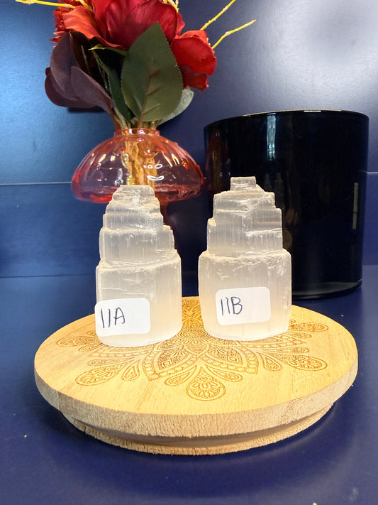 Two translucent white selenite towers on a decorative wooden coaster in front of a red flower and black jar