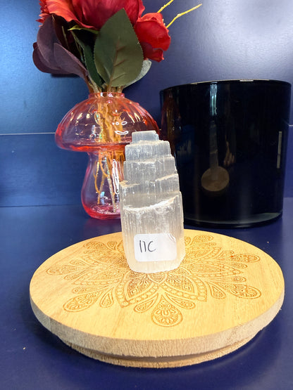 Two translucent white selenite towers on a decorative wooden coaster in front of a red flower and black jar