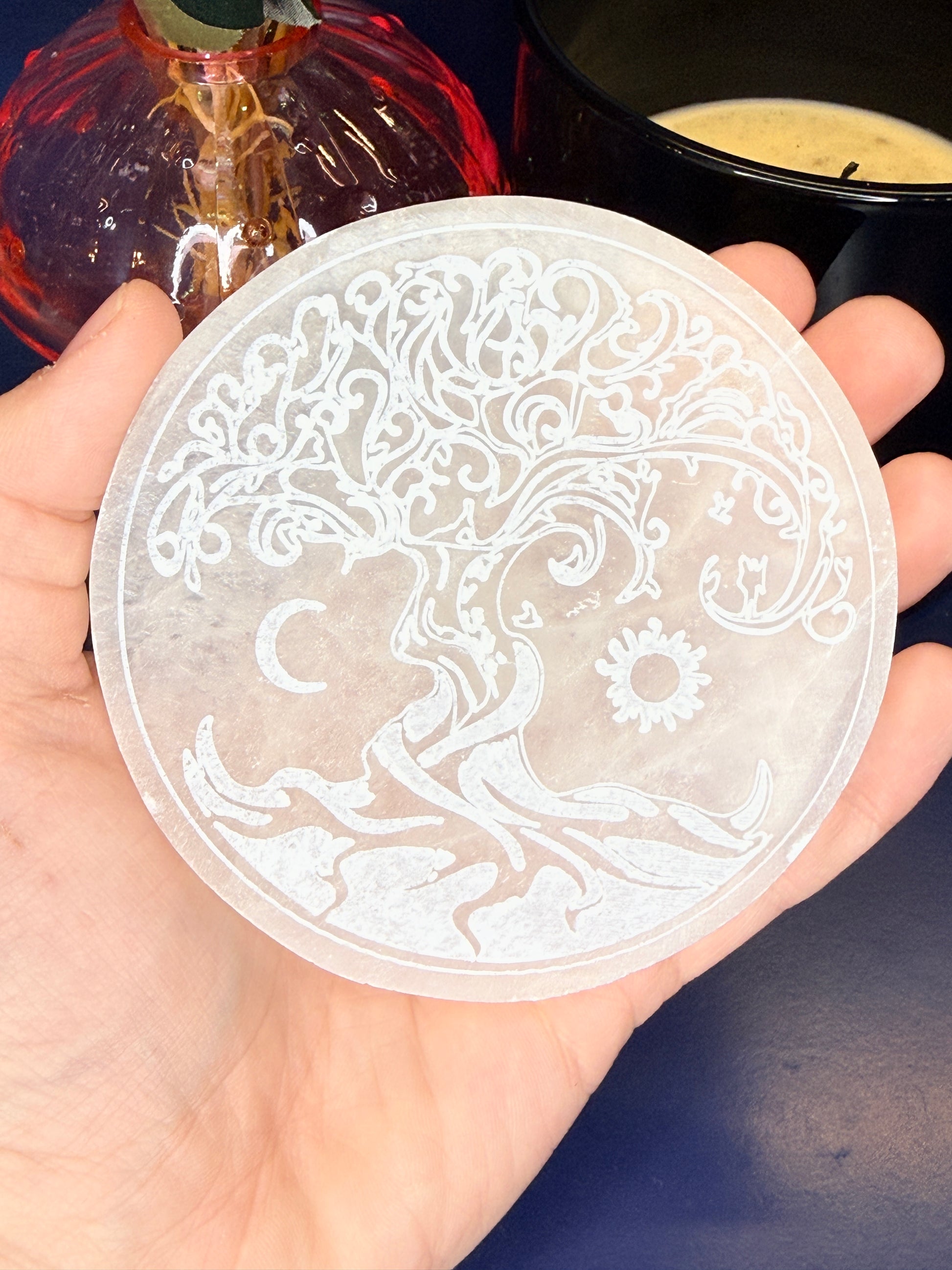 Hand holding a round tree of life selenite charging plate in front of a pink mushroom vase and black candle jar