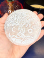 Hand holding a round tree of life selenite charging plate in front of a pink mushroom vase and black candle jar