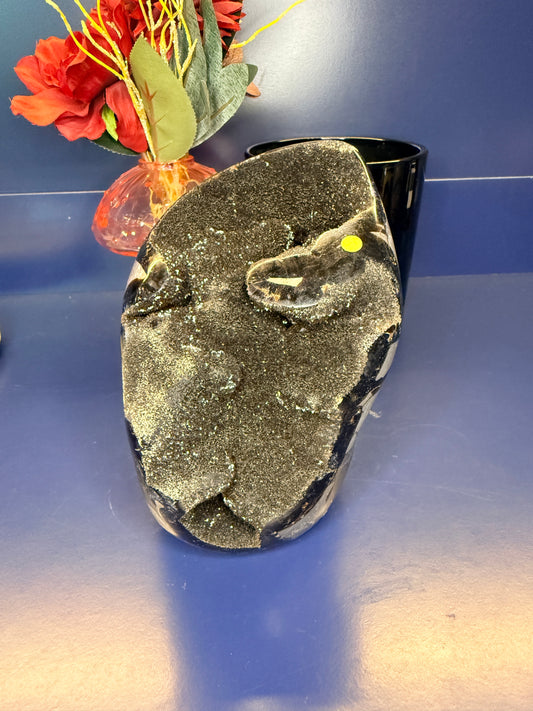 Black septarian geode freeform in front of a red flower in a vase and black jar on a blue background