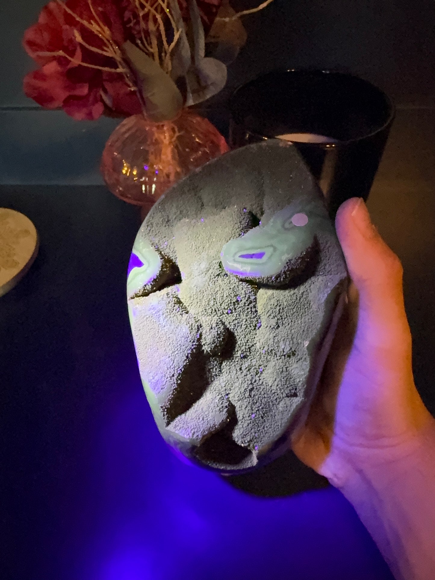Hand holding a glowing green and purple septarian geode freeform in front of a red flower in a vase and black jar