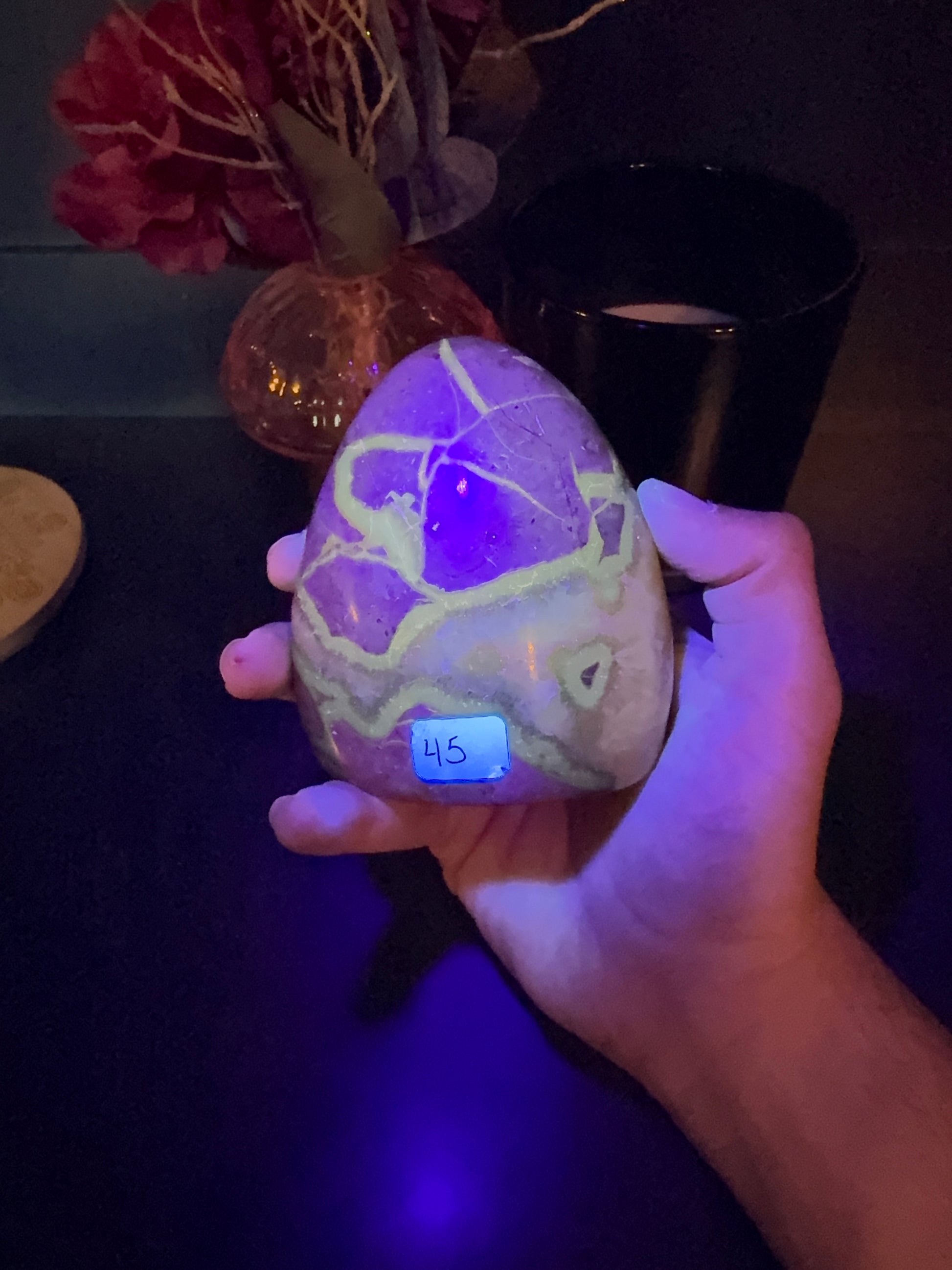 Hand holding a glowing green septarian freeform in front of a red flower in a vase and black jar
