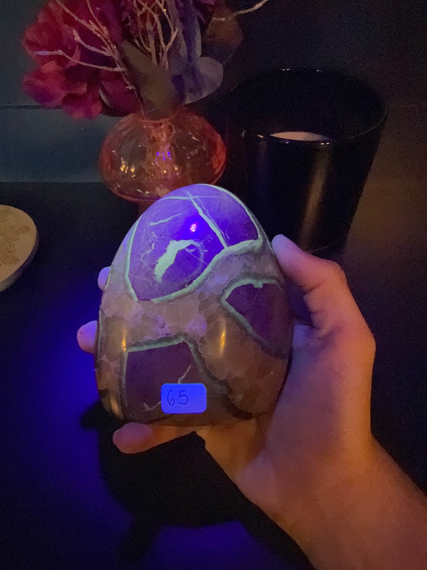 Hand holding a glowing green septarian freeform in front of a red flower in a vase and black jar