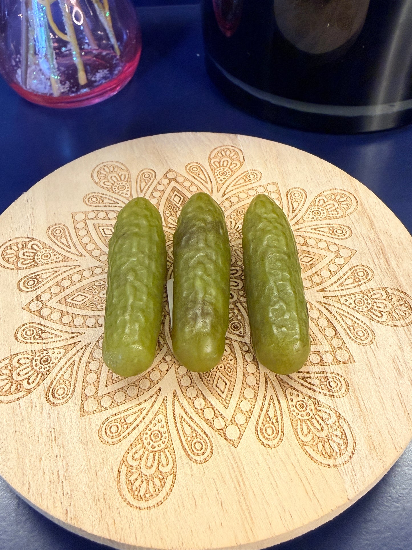 Three green crystal pickles sitting on a decorative wooden coaster in front of a glass mushroom and black jar