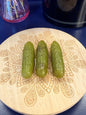 Three green crystal pickles sitting on a decorative wooden coaster in front of a glass mushroom and black jar