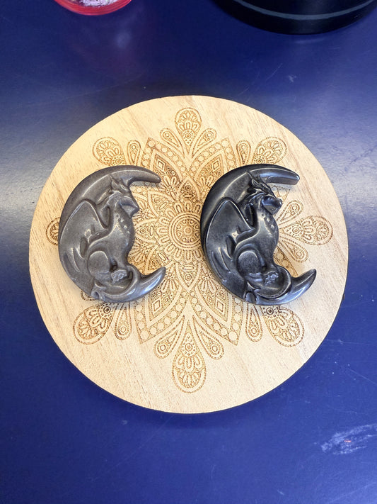 Two silver and black obsidian dragons in moons on a decorative wooden coaster against a blue background