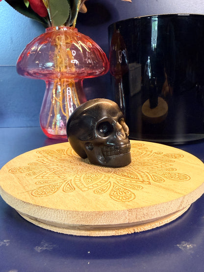 Black and silver obsidian skull shape sitting on a decorative wooden coaster in front of a mushroom and black jar against a blue background