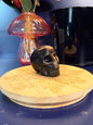 Black and silver obsidian skull shape sitting on a decorative wooden coaster in front of a mushroom and black jar against a blue background