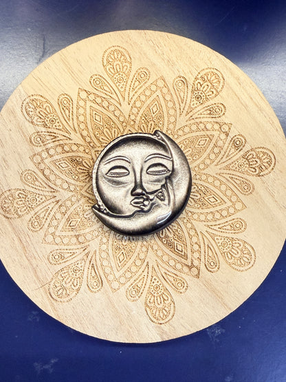 Silver and black obsidian sun & moon shape sitting on a decorative wooden coaster against a blue background