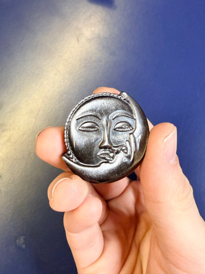 Hand holding a silver and black obsidian sun & moon shape sitting against a blue background