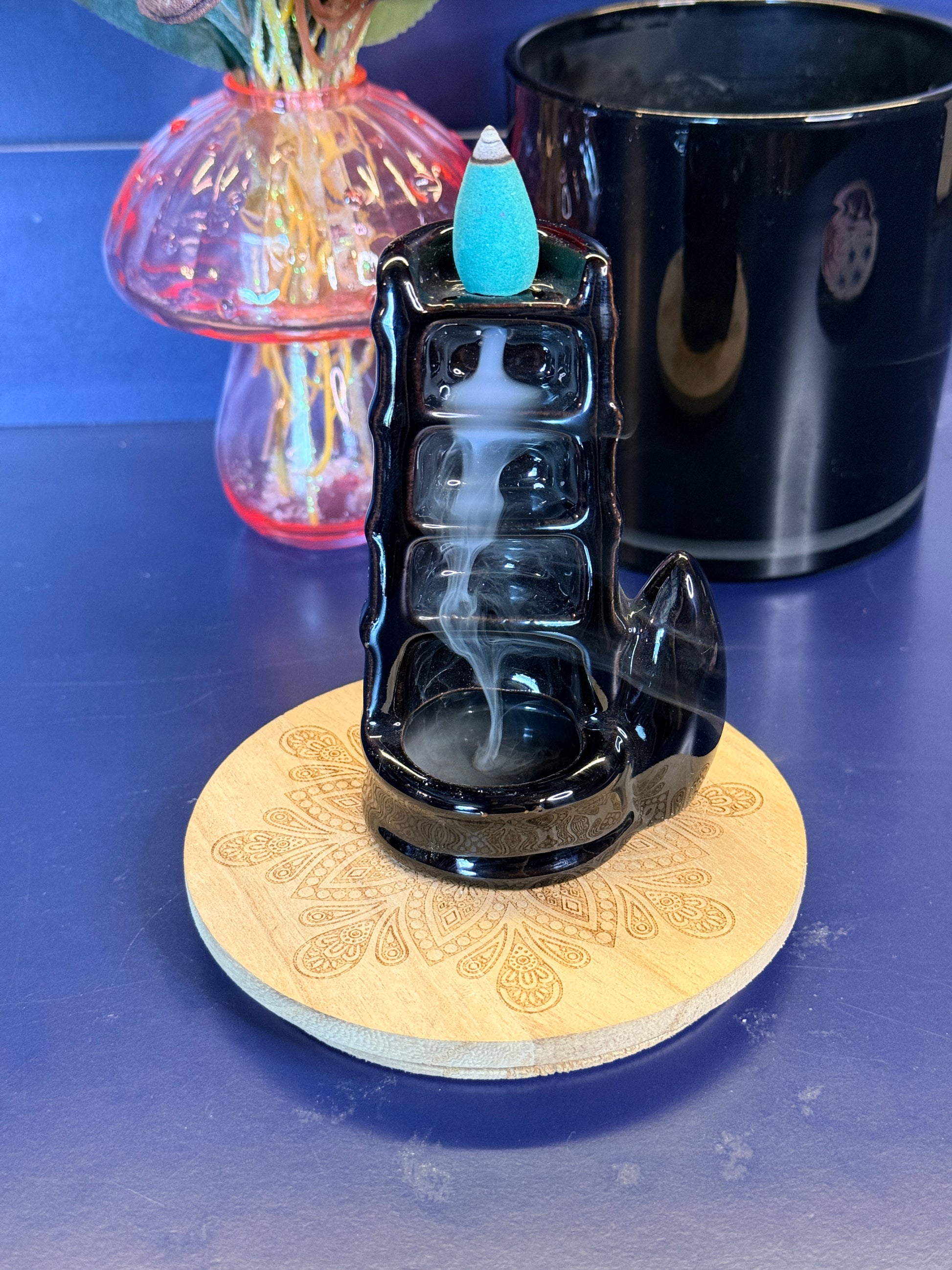 Black waterfall incense burner with a blue incense cone on top, in front of a pink mushroom and black jar on a blue background