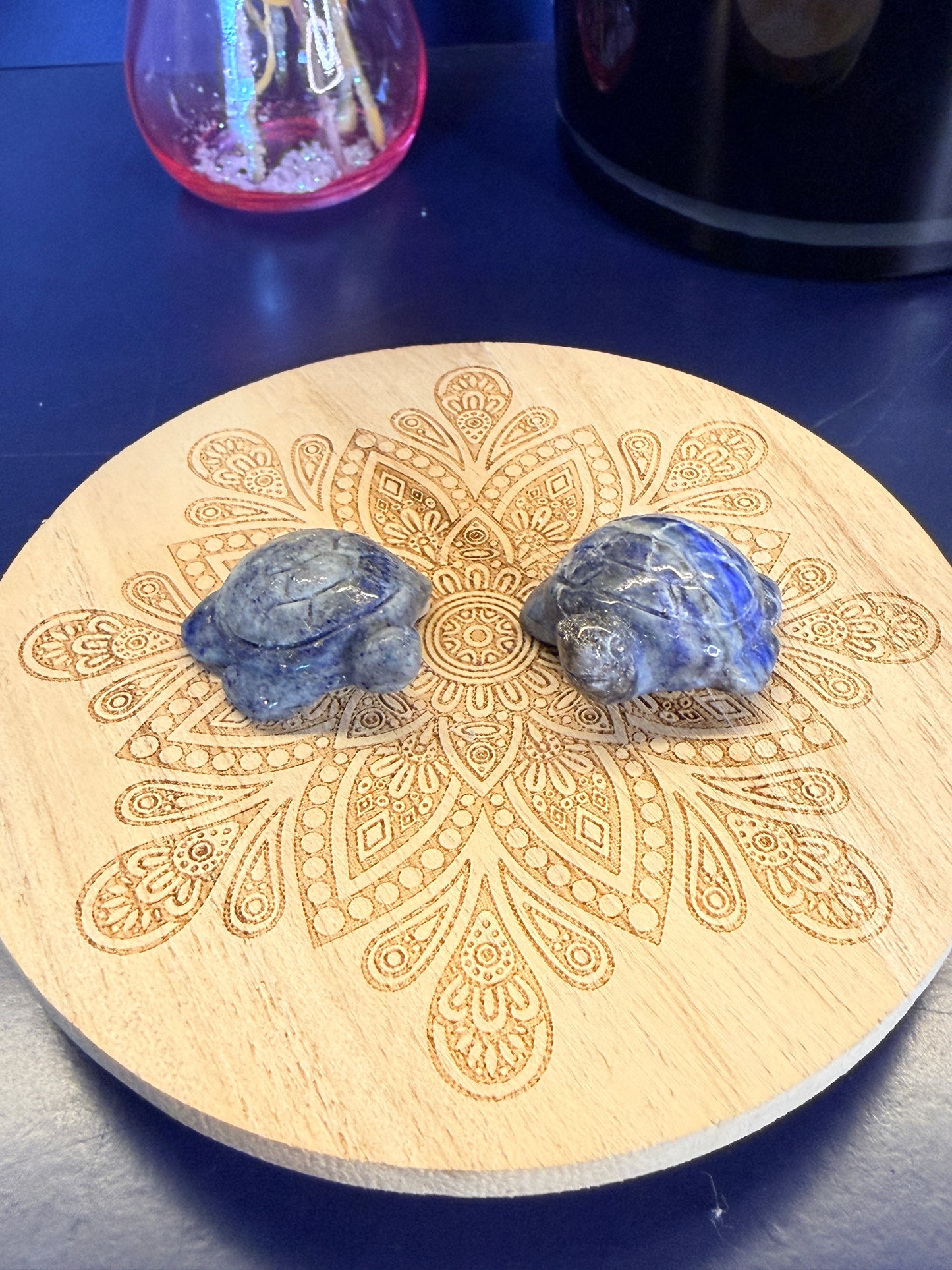 Two Blue and grey crystal turtles on a decorative wooden coaster in front of a vase and black jar